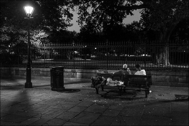 two men sitting on park bench at night; one man sleeping on seat behind them
