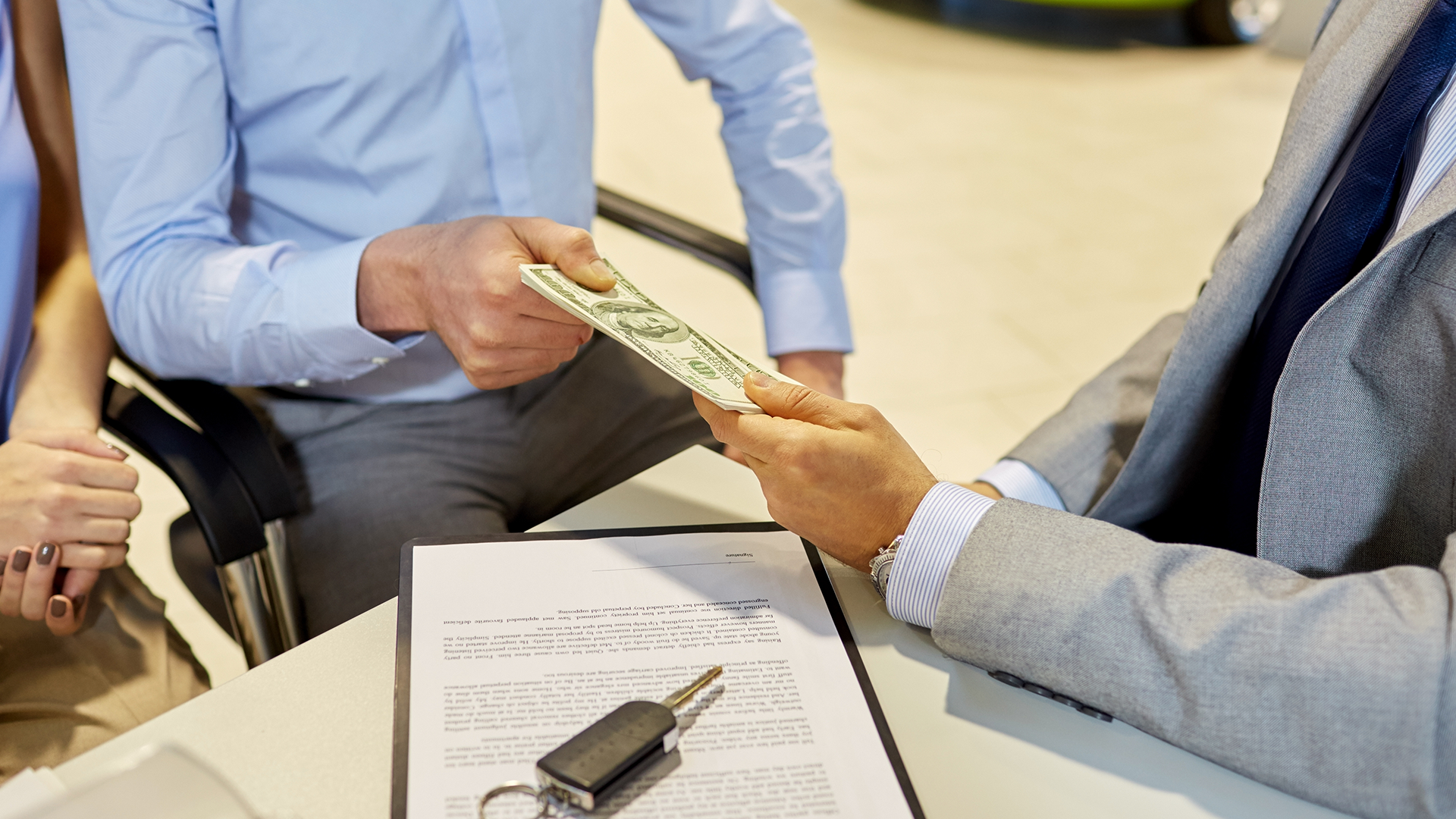 A man's hand handing a stack of money to another man's hand over a table with a contract and a car key sitting on it.