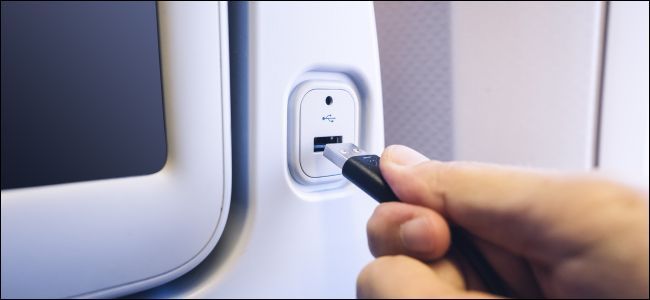 A hand plugging a USB cord into a charging port on the back of an airline seat.