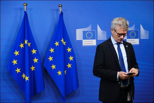 A man using a smartphone with two EU flags behind him.
