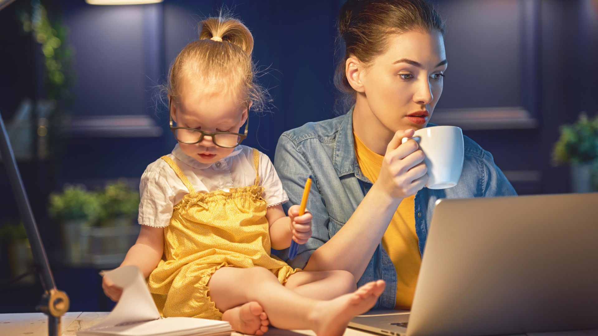 A woman with a laptop and coffee trying to get work done with a toddler sitting beside her