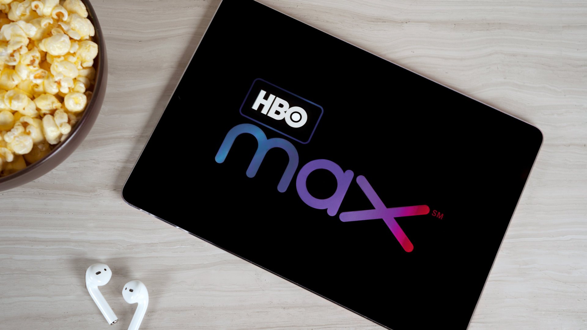 HBO Max displayed on a tablet next to a pair of AirPods and a bowl of popcorn