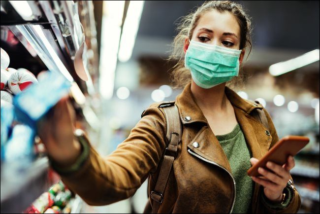A woman wearing a face mask and holding a phone while taking something off a store shelf. 