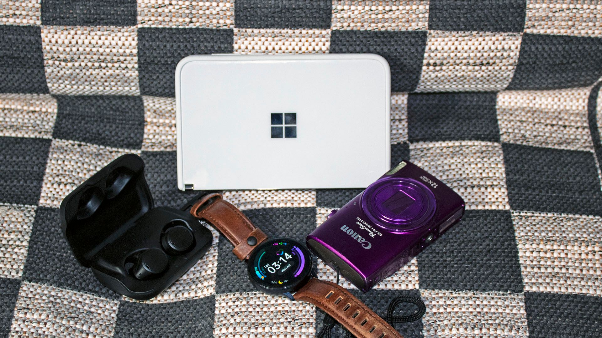 A Surface Duo, smartwatch, handheld camera, and true wireless earbuds on a checkered blanket.
