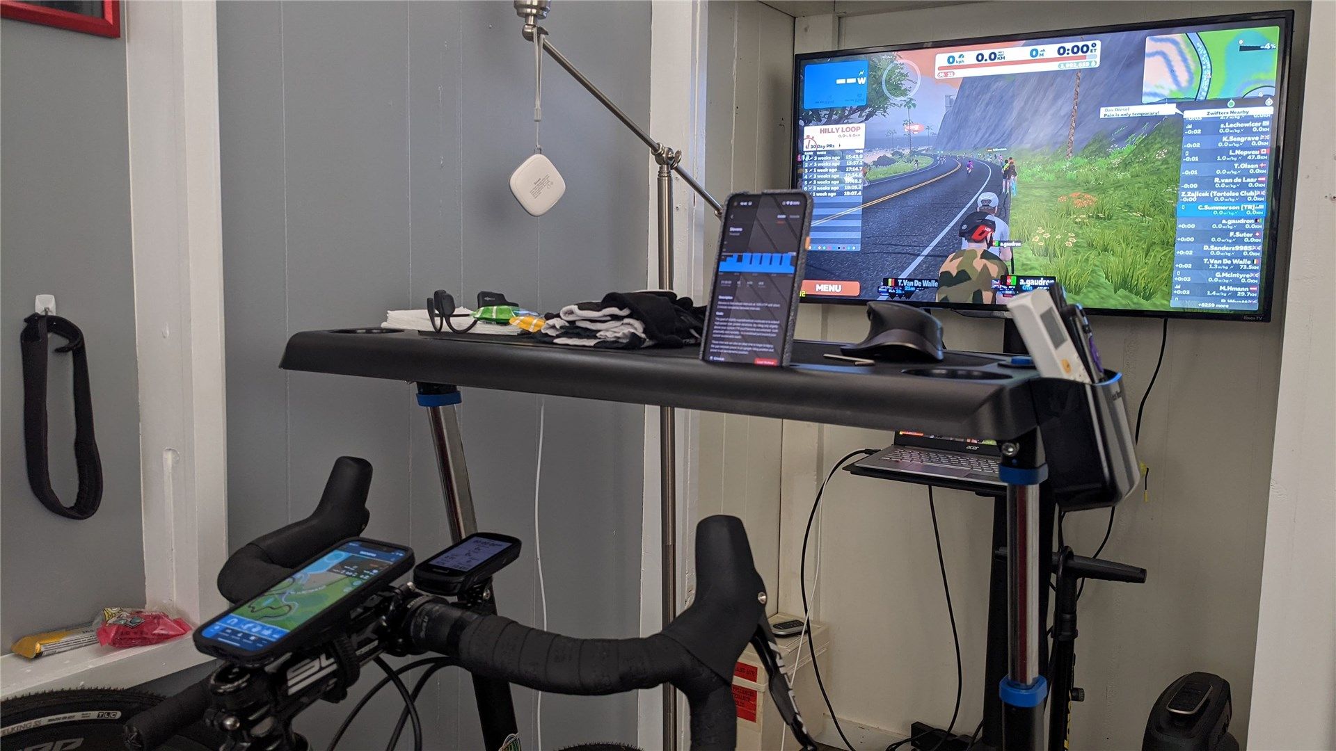 An indoor training setup with Zwift on a TV, TrainerRoad on a phone, and Gearlock holding an iPhone with the Zwift companion app.