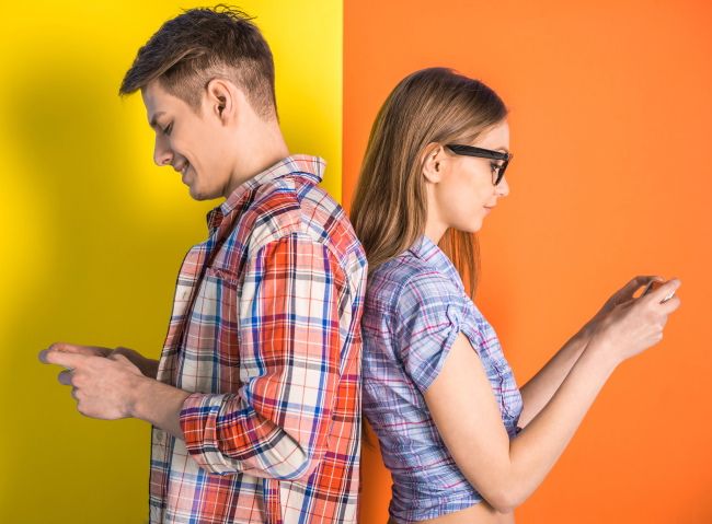 A young couple standing back to back while using their phones.