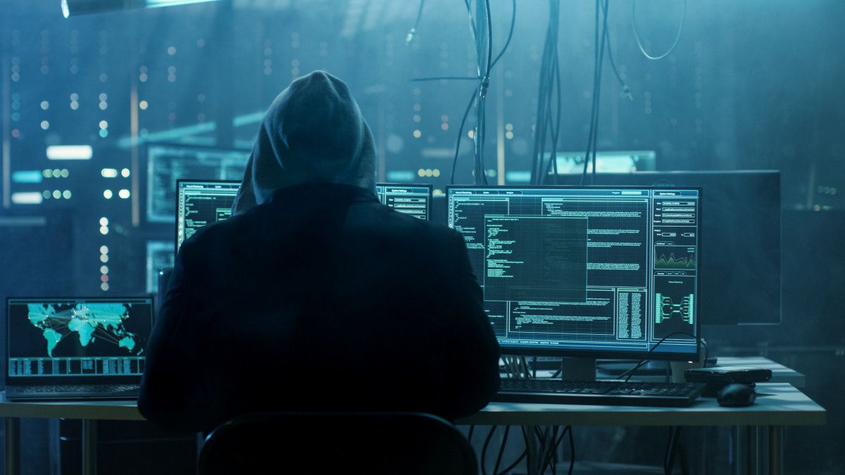 A hooded hacker sitting in front of computers.