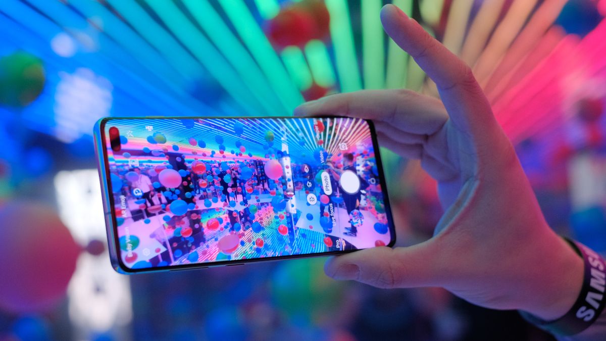 Hand holding a Samsung Galaxy S10 with a coloful display in front of multicolored lights.