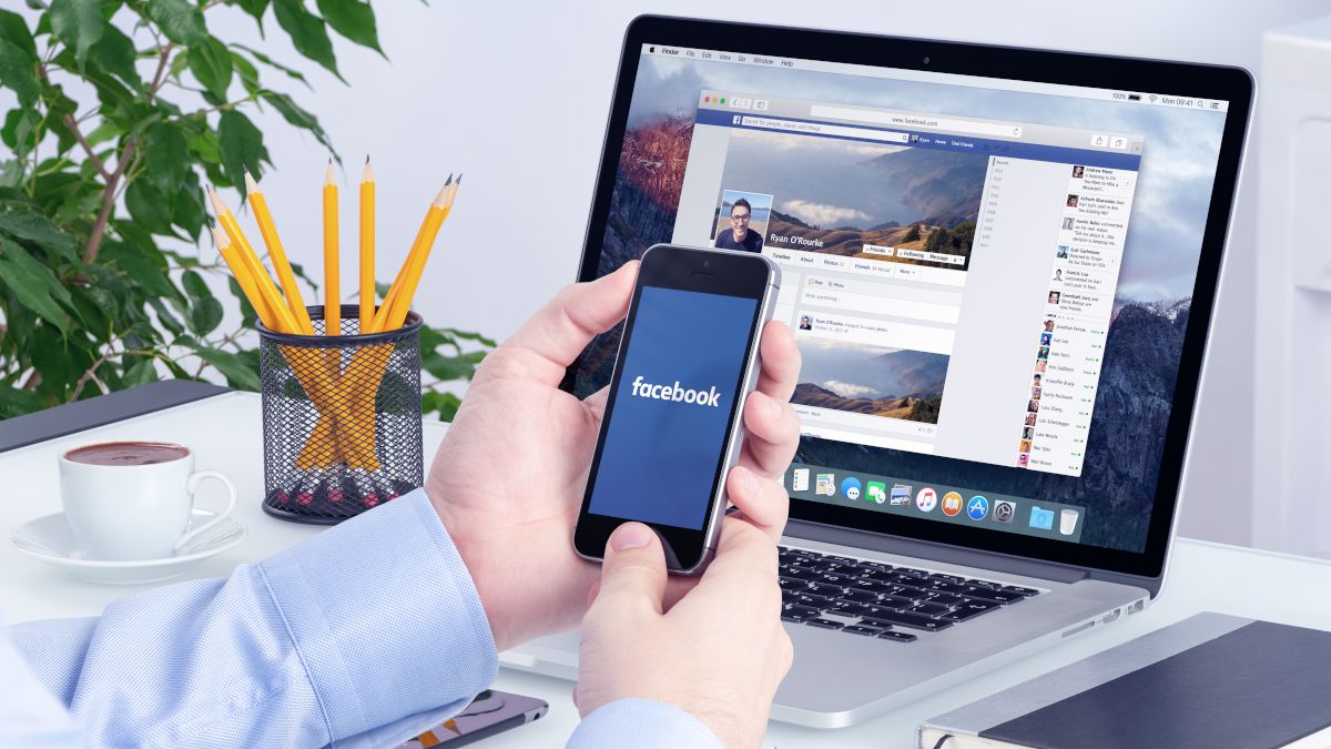 Hands holding a smartphone in front of laptop with Facebook open