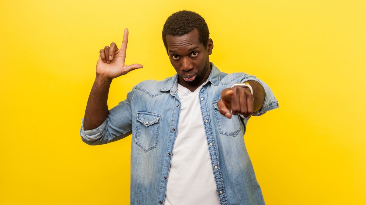 Man pointing with loser hand gesture on yellow backdrop