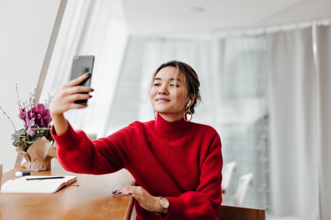 Woman in a red sweater taking a selfie in the natural light of a window
