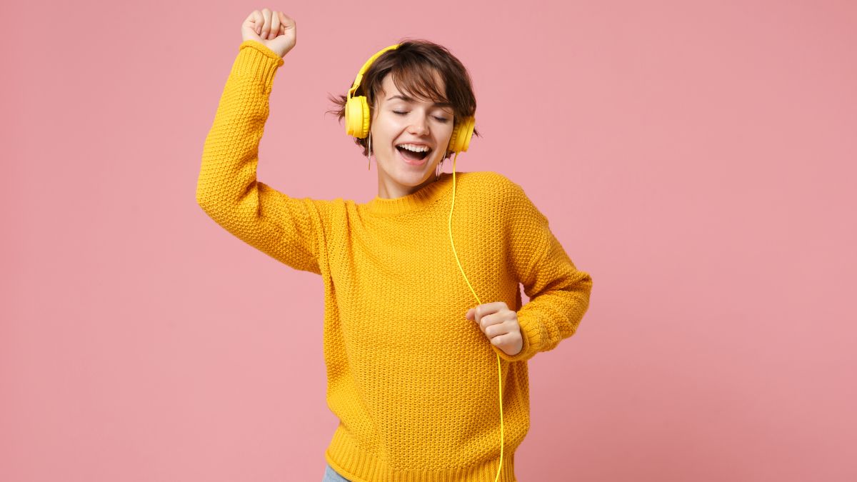 Woman in yellow sweater dancing while listening to music on yellow, wired headphones