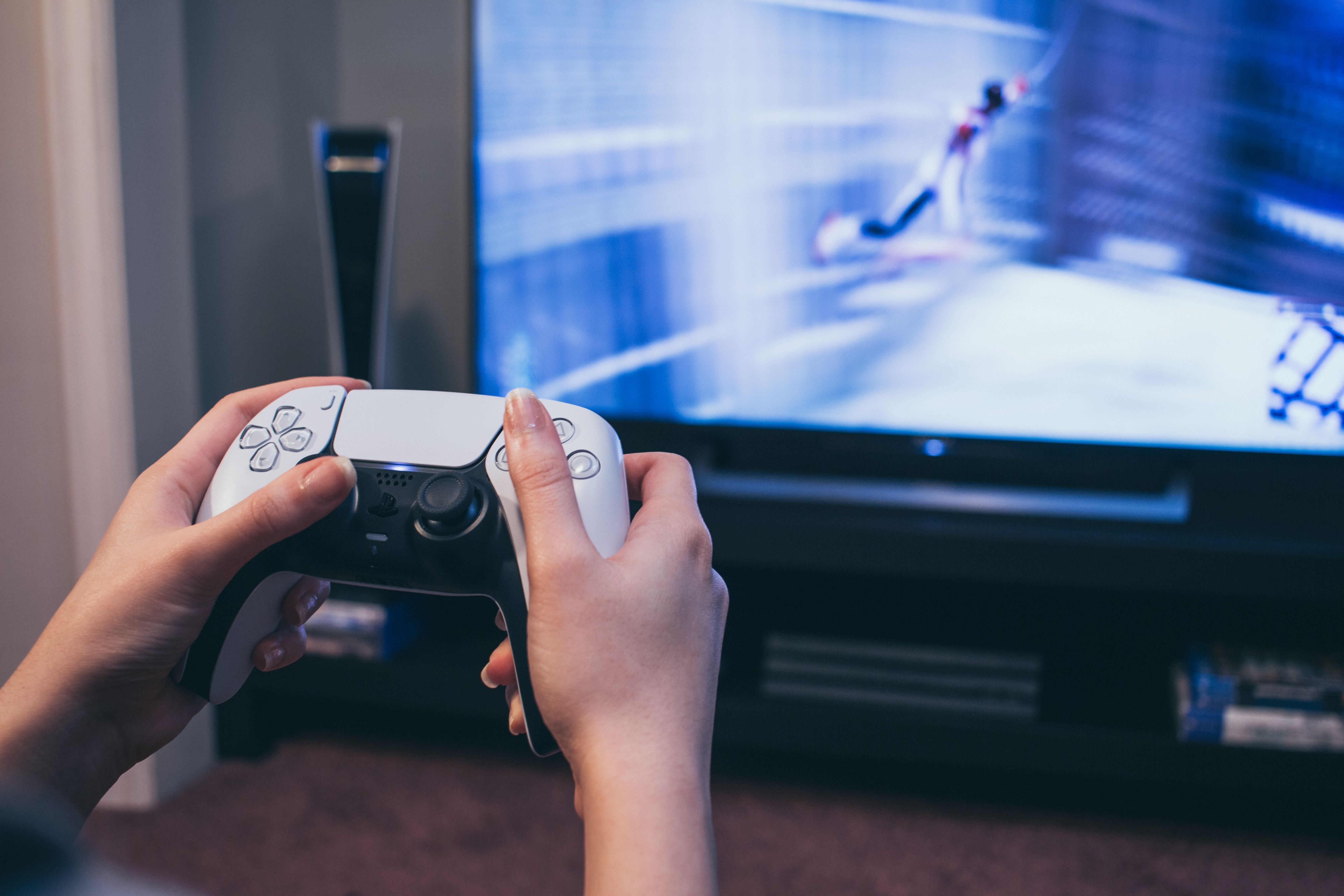 Hands holding a PlayStation 5 controller in front of a TV.