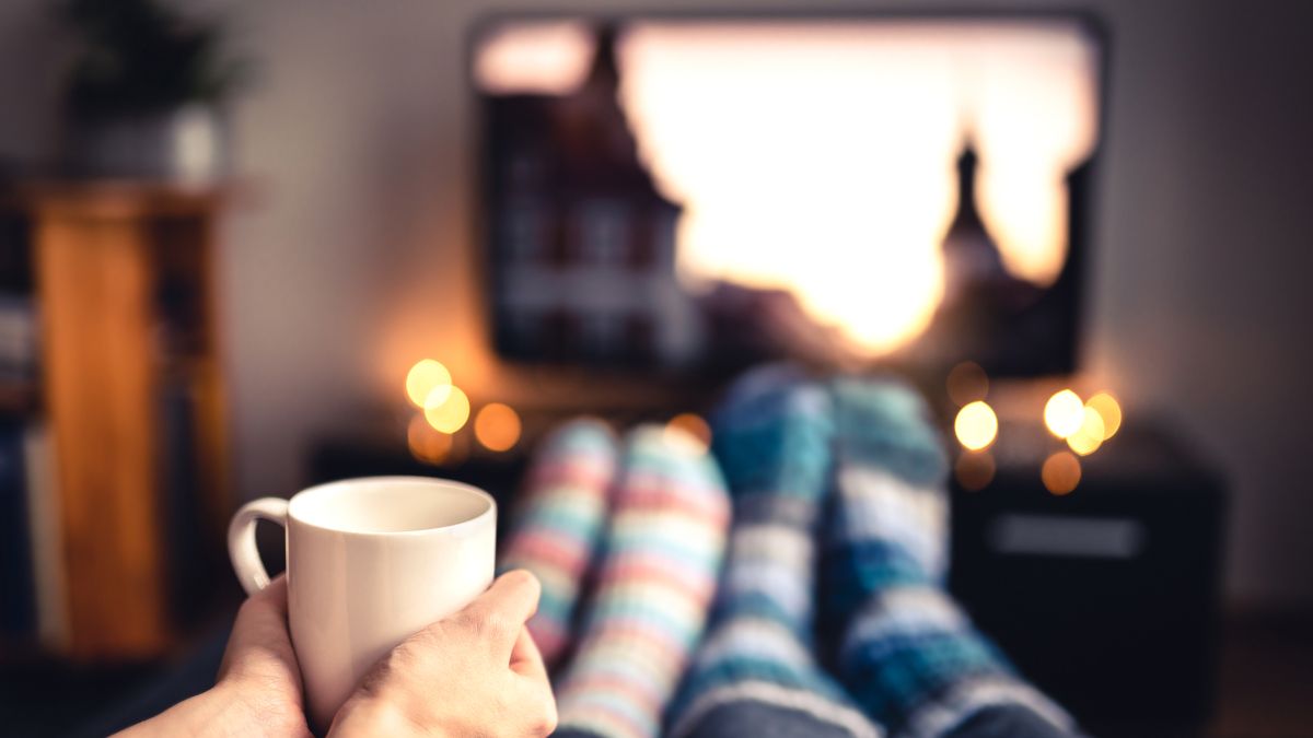 Point-of-view shot of a couple drinking hot chocolate and watching television with Christmas lights in view.