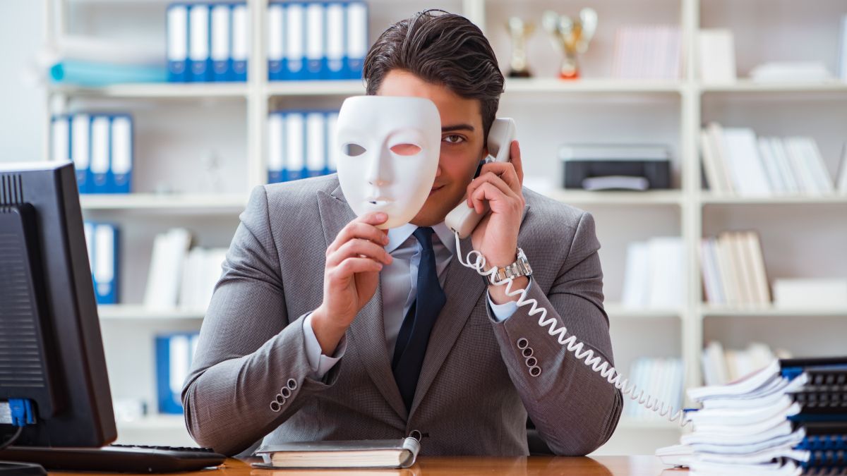 A businessman in an office making a phone call and peeking out from behind a face mask.