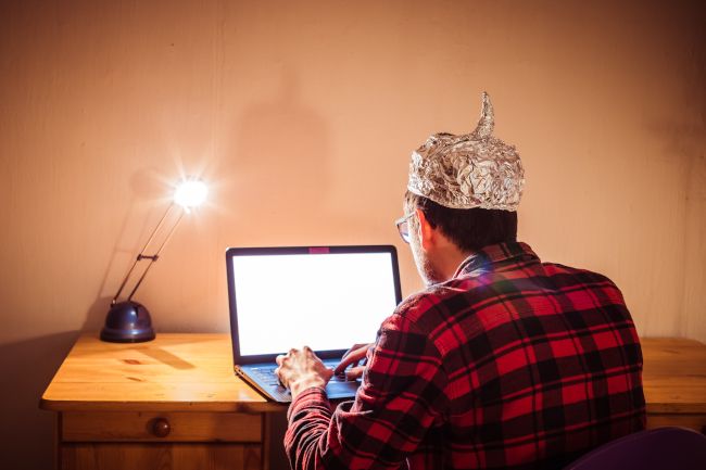 Young man sitting at a desk with a laptop and a tin foil hat.