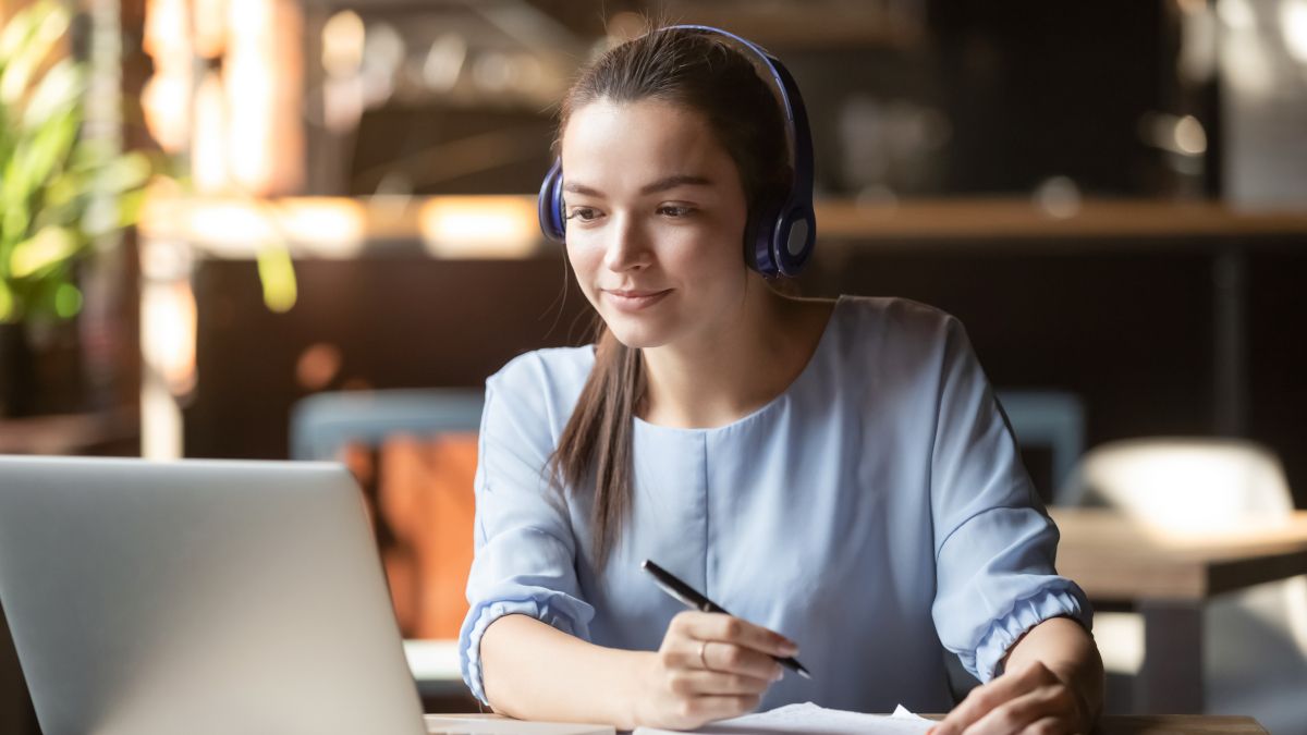 Young woman wearing headphones while taking notes in front of a laptop,