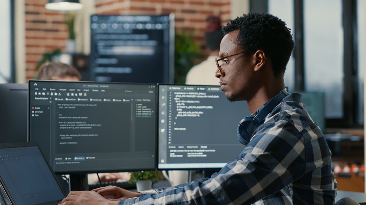 You man using a laptop next to multiple computer monitors with code on display.