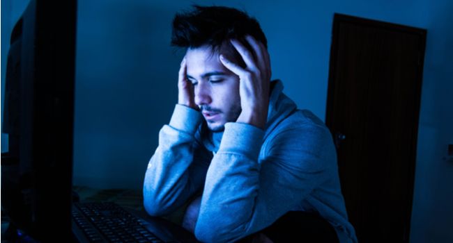 Man looking stressed or exhausted while using a computer in the dark.