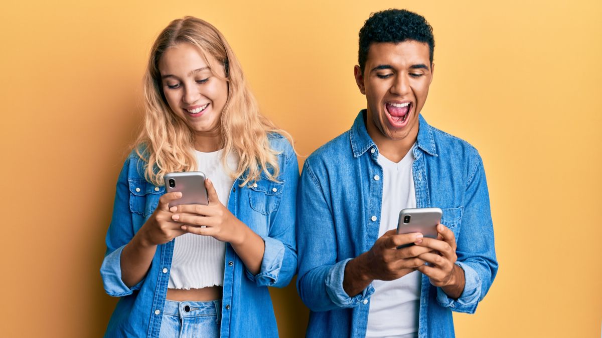 A young couple standing next to each other while smiling and using their smartphones.