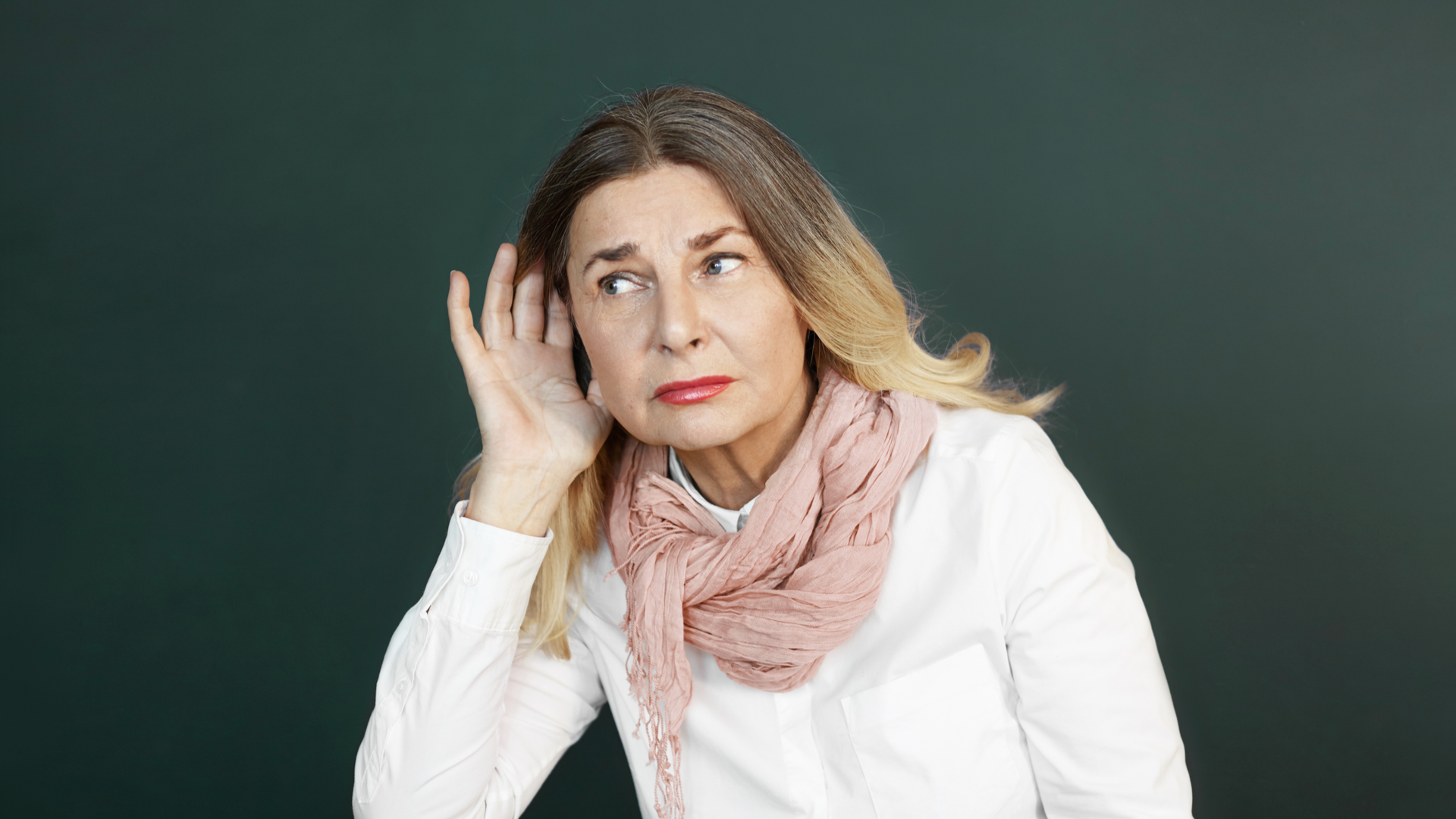 Studio shot of stylishly dressed beautiful retired female with blonde hair holding hand at her ear while overhearing interesting conversation, struggling to hear some news, secret or gossip