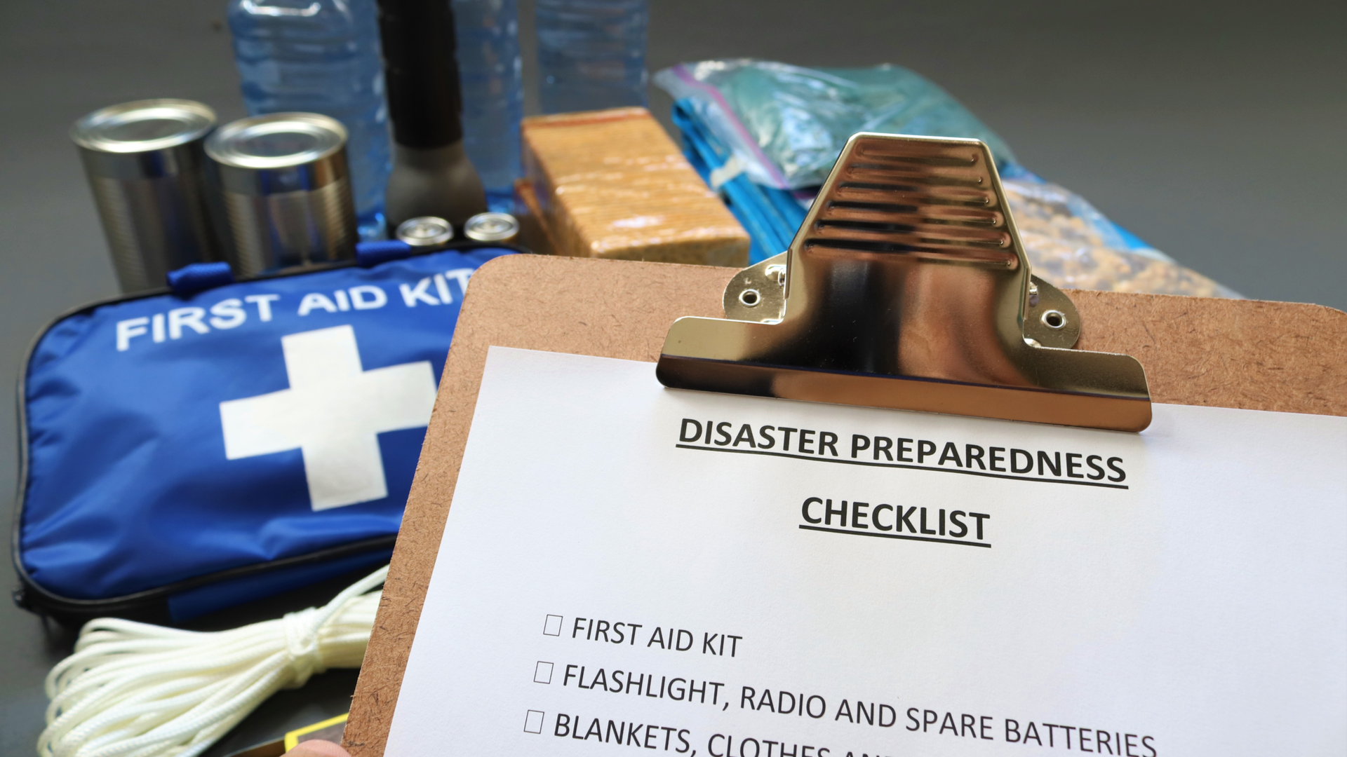 Disaster preparedness checklist on a clipboard with disaster relief items in the background.Such items would include a first aid kit, flashlight, tinned food, water, batteries and shelter.