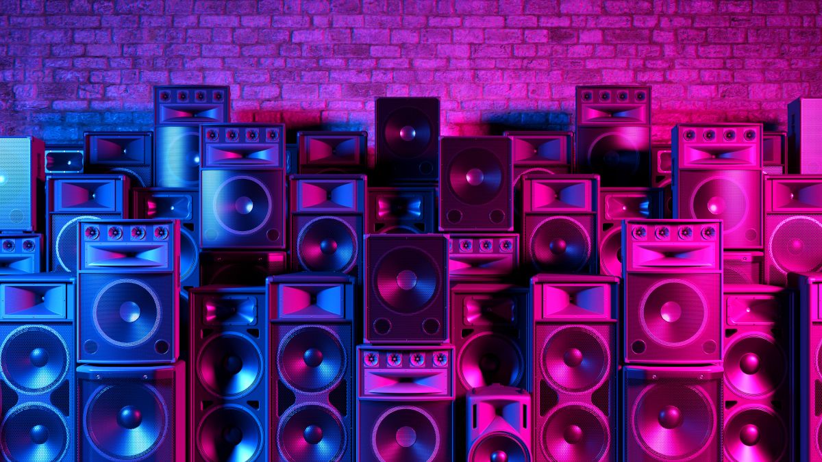 A large collection of speakers standing against a brick wall in blue and pink lighting.