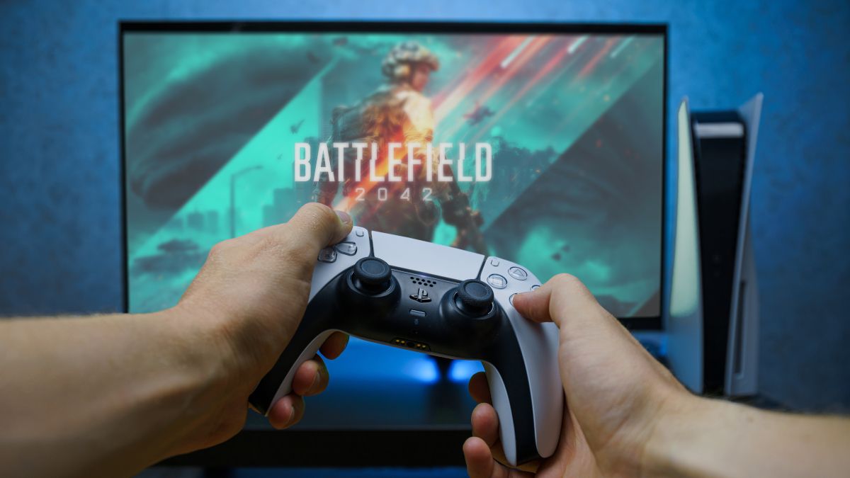 Hands holding a PlayStation 5 controller in front of a computer monitor, playing Battlefield 2042.