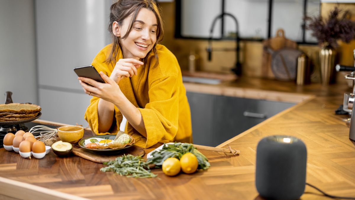 Woman in a kitchen using voice commands to control a smart speaker.