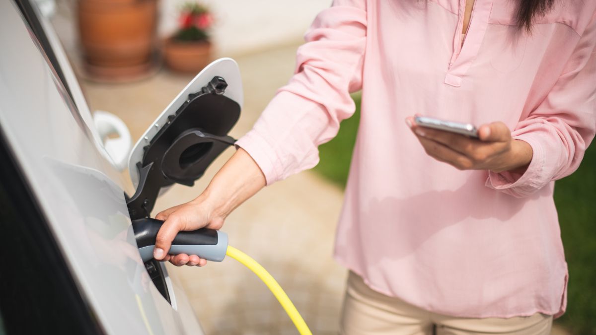 Woman charging an electric car while looking at a smartphone.