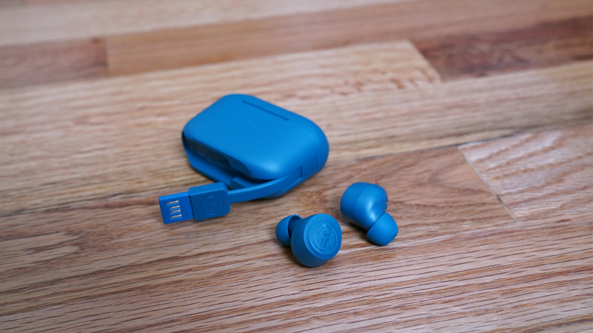 The JLAB Go Air Pro earbuds laying on a wooden floor next to their case, showing the attached USB-A charging cable