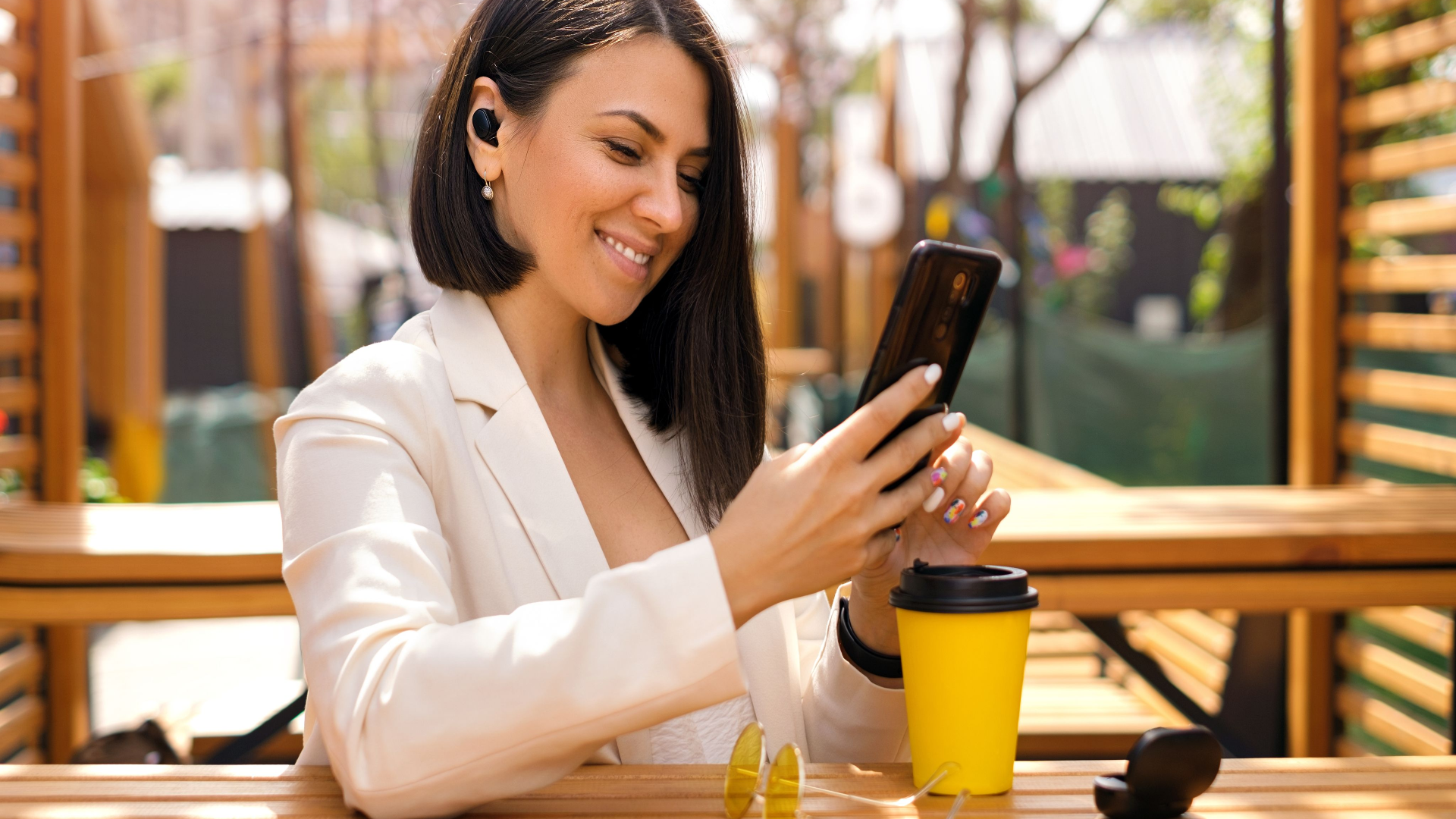 Brunette business person with small wireless black headphones in ears looks at the phone with a smile, reading messages. Person is relaxing in a cafe on the terrace enjoying music