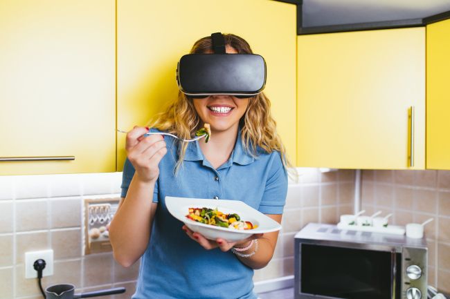 Woman in a kitchen wearing a virtual reality headset and eating a bowl of food.