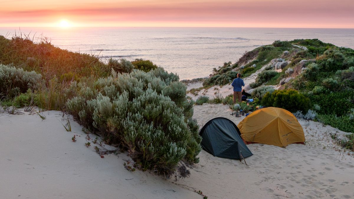 Person on the beach of Margaret River in Australia, standing next to two tents and looking at the sunset.
