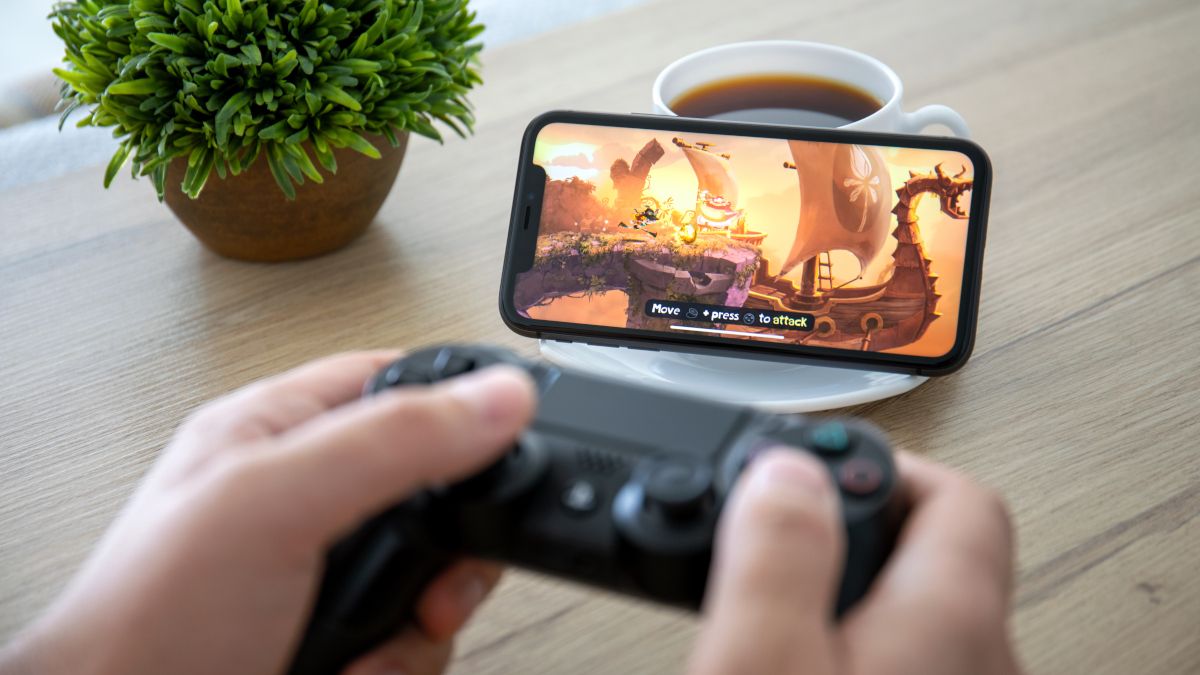 Person holding a DualShock 4 controller in front of an iPhone 11 playing Rayman Adventures.