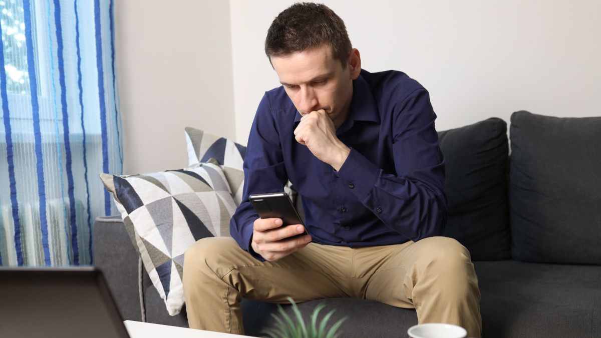 A young man sitting on a couch and looking at his phone with a worried expression.