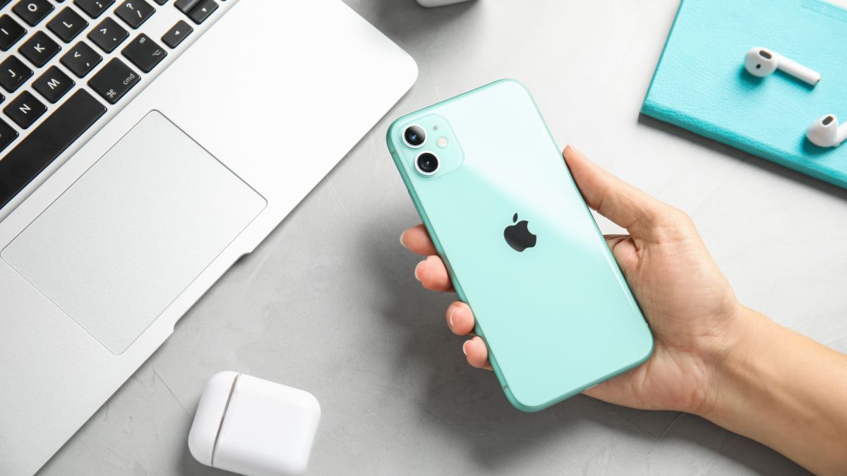 A woman's hand holding a green iPhone 11 with AirPods and a MacBook visible in the background.