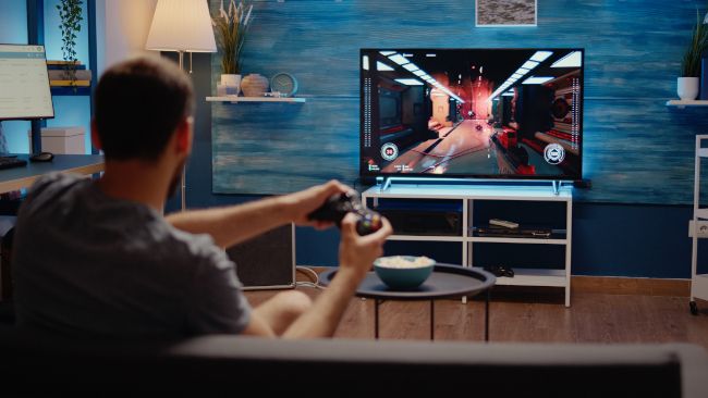 Over-the-shoulder view of a man playing an FPS game on a TV.