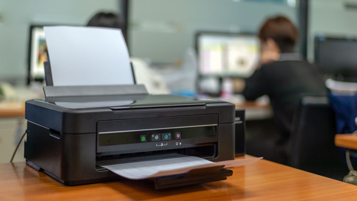 An office printer on a desk with a person in the background using a computer.