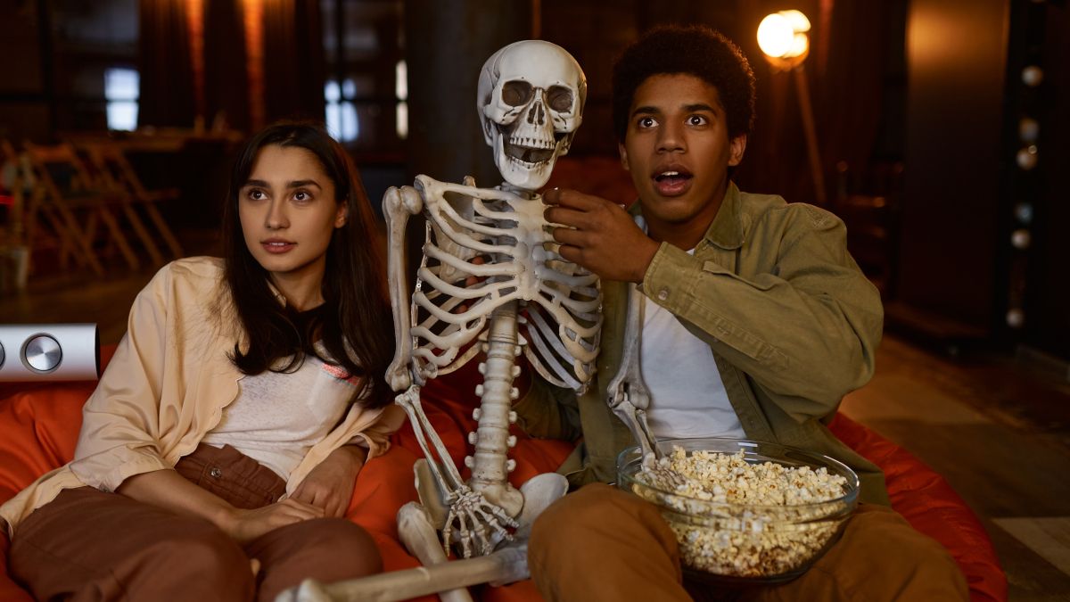 A young couple sitting on a couch with a model skeleton and watching a movie.