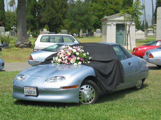 Photo of several EV1s parked in a cemetery, with flowers and a drape covering one of the cars