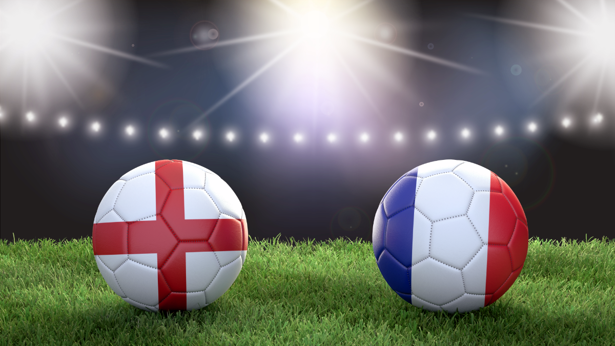 Two soccer balls emblazoned with the colors of the English and French flags sit on a soccer field underneath stadium light