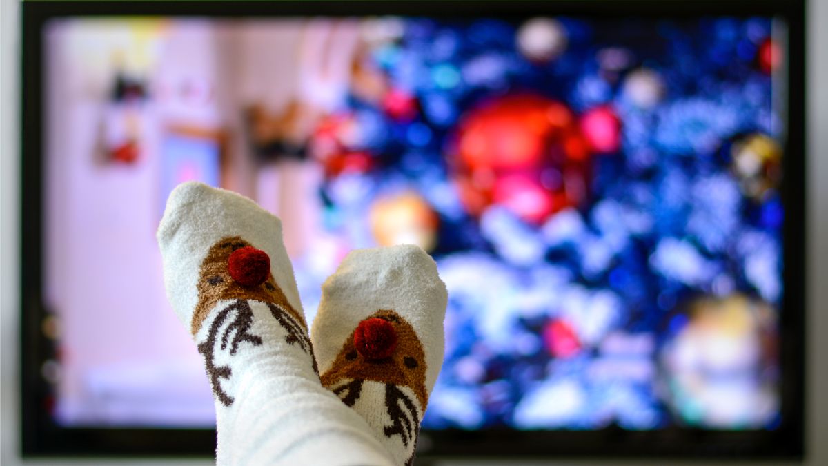 Point of view shot of a person's socks with reindeer designs and a Christmas movie on a TV screen in the background.