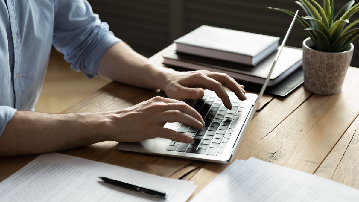 Person's hands typing on a laptop with paperwork and notebooks on the desk.
