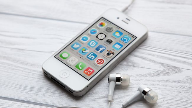 An iPhone 4 with earbuds on a white wood tabletop.