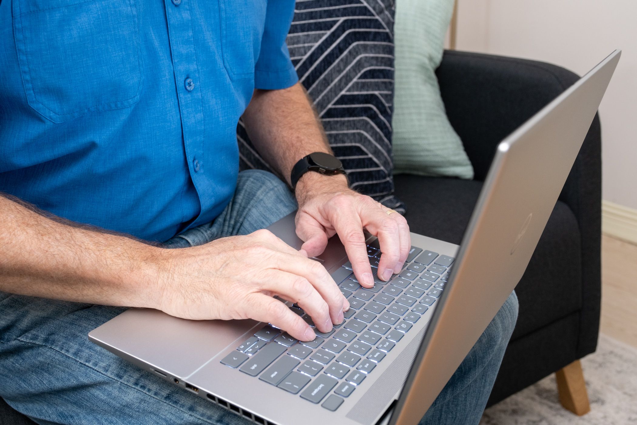 A man sitting on a couch is typing on a silver laptop.
