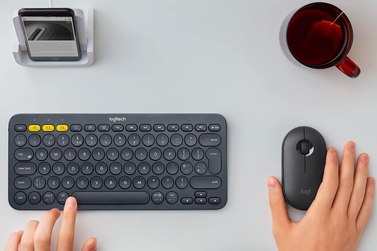 A Logitech K380 on a desk with a person's hands, one on the keyboard and one on a mouse.