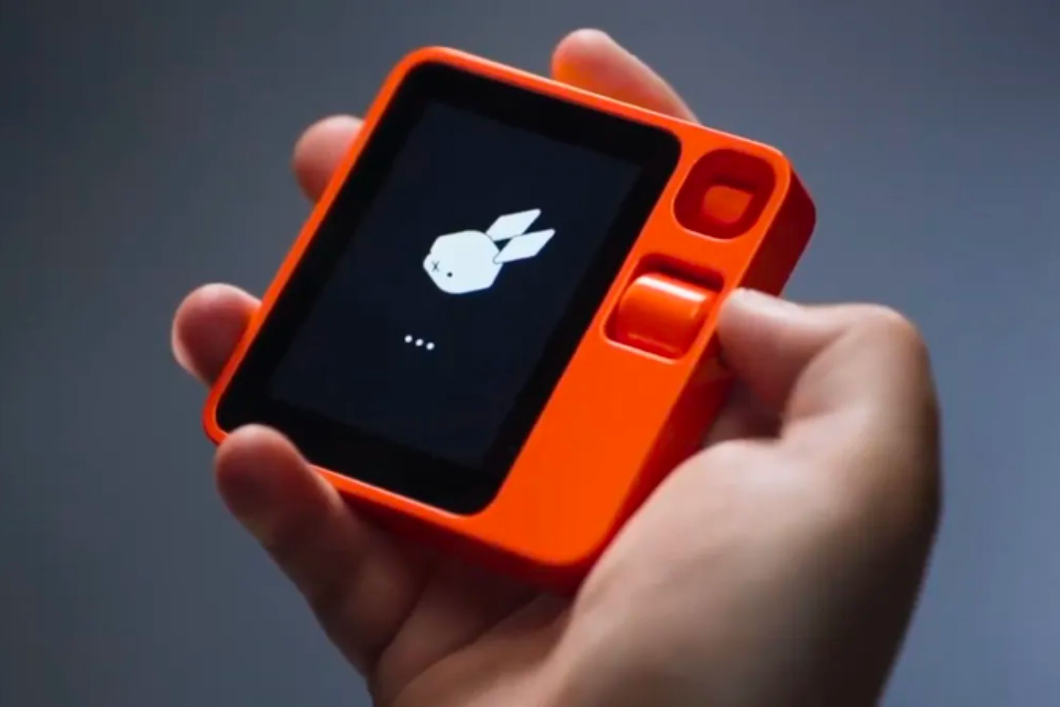 The R1 Rabbit companion device held in a hand, showing the Rabbit icon on its screen.