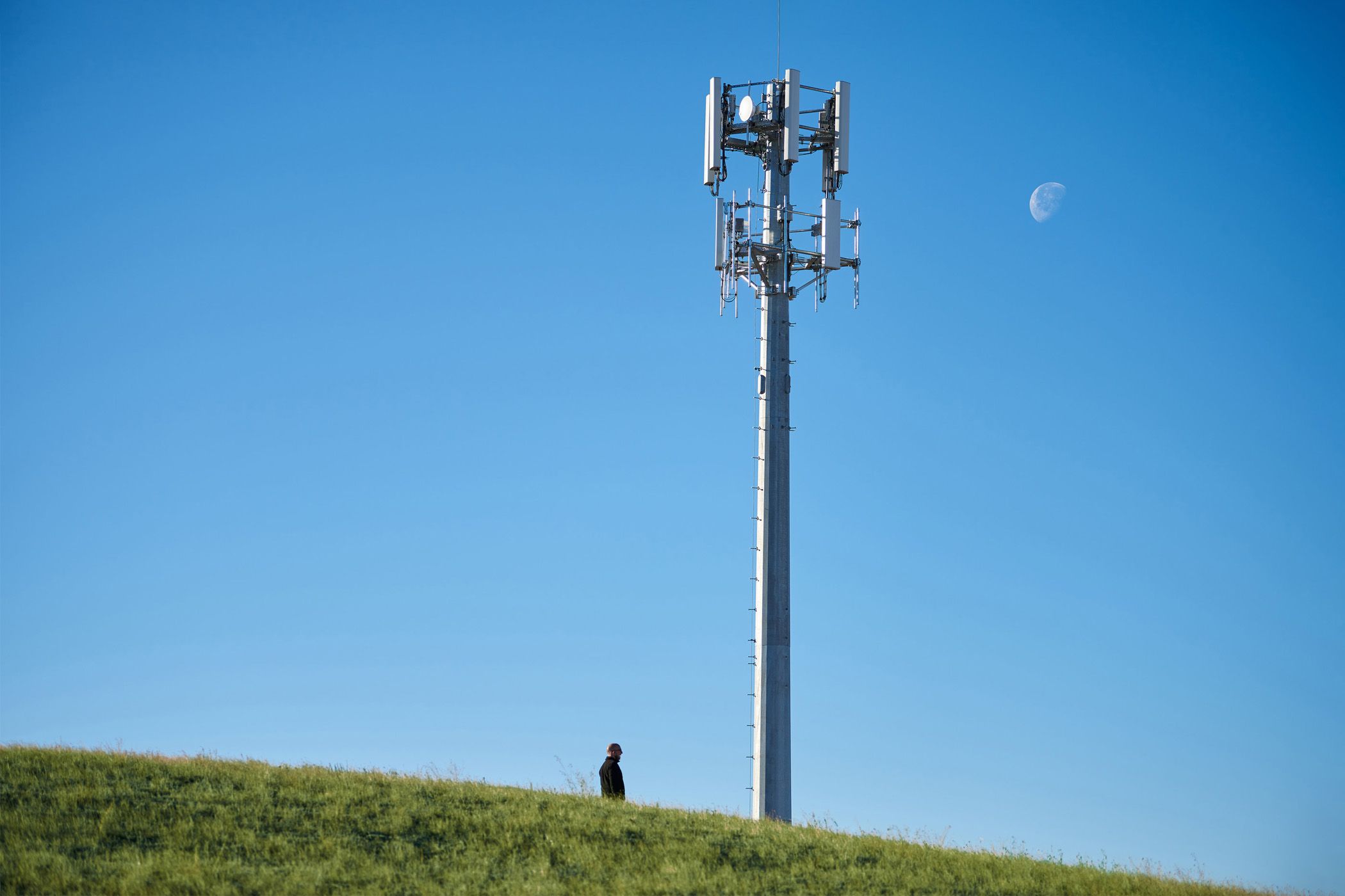 Cell tower on a hill with a person standing below and a daytime moon in the sky.