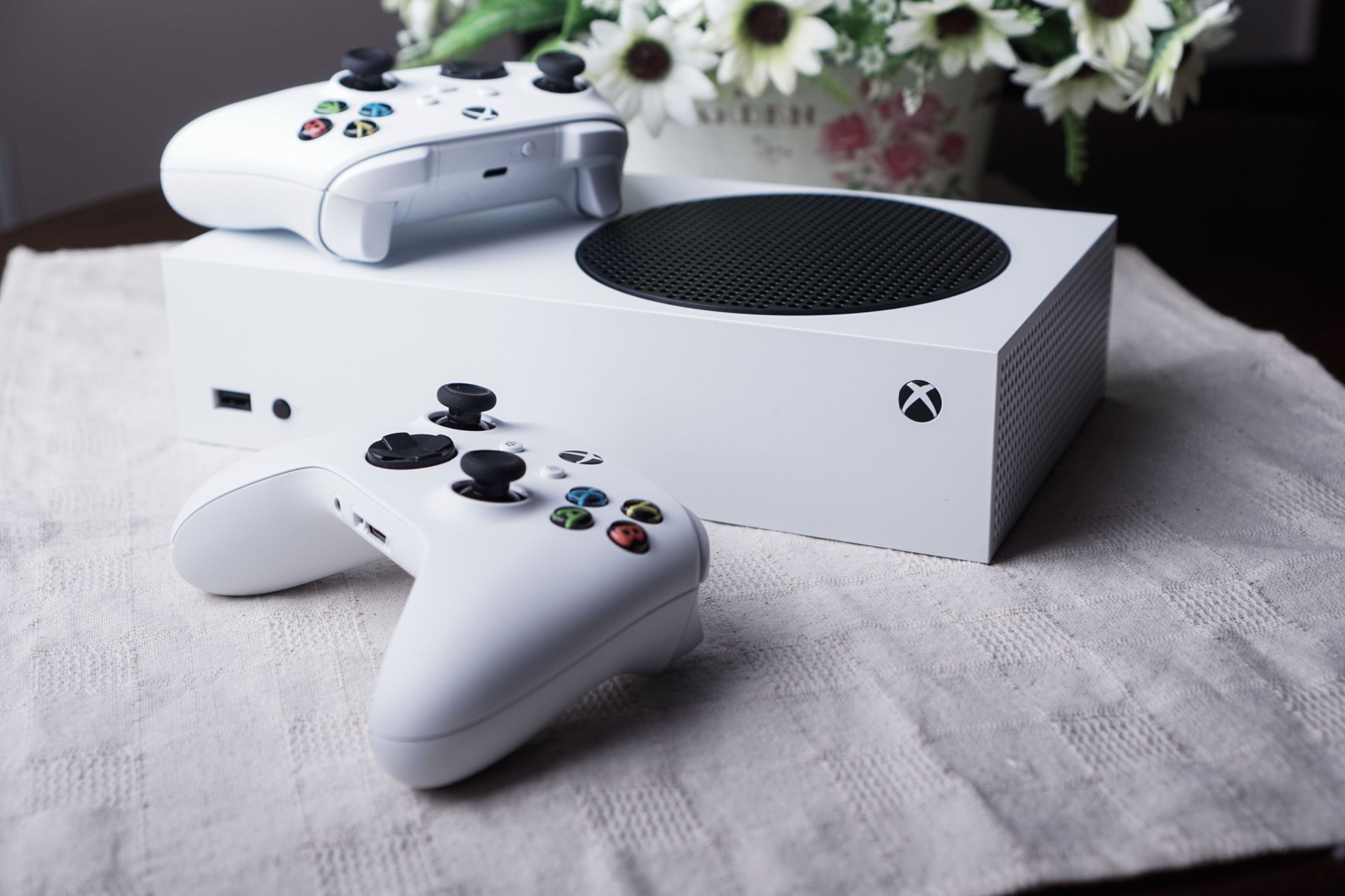 A Series S console on a table with two controllers and a vase of flowers.